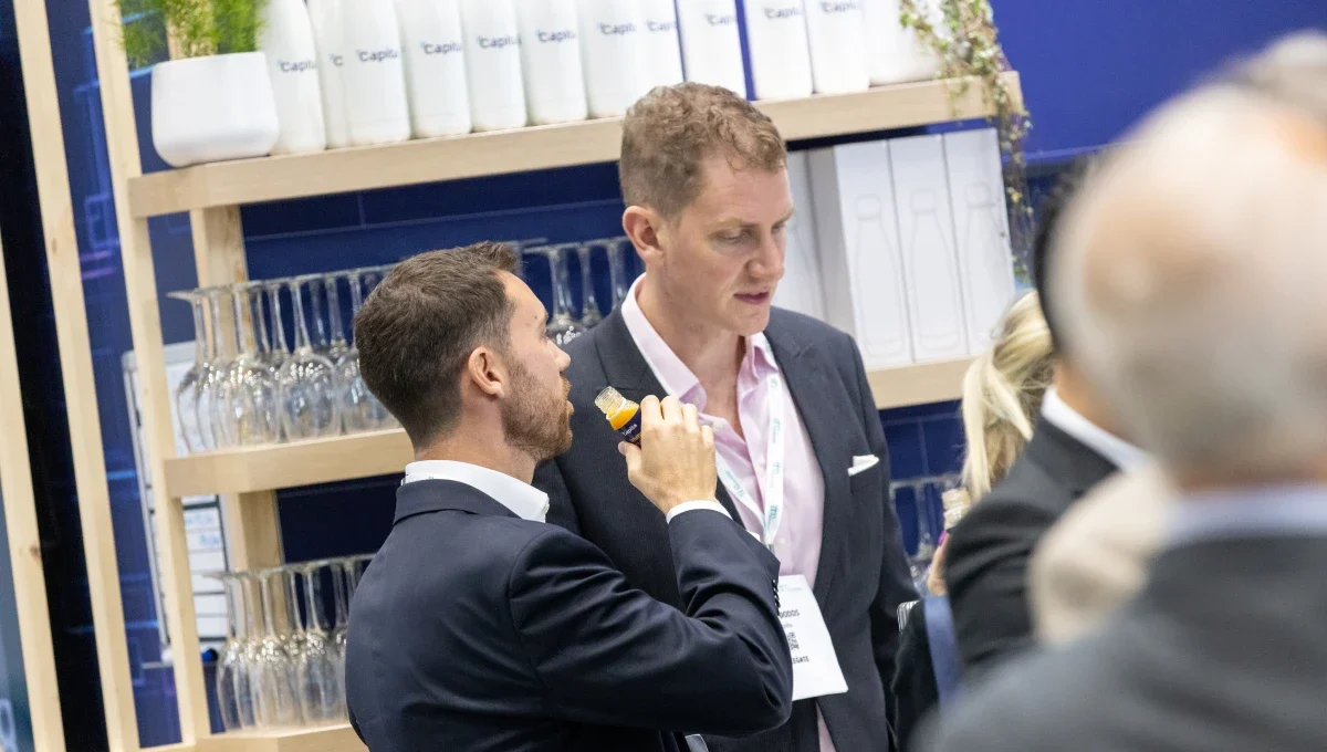 Two men speaking at an event with one man drinking a juice shot